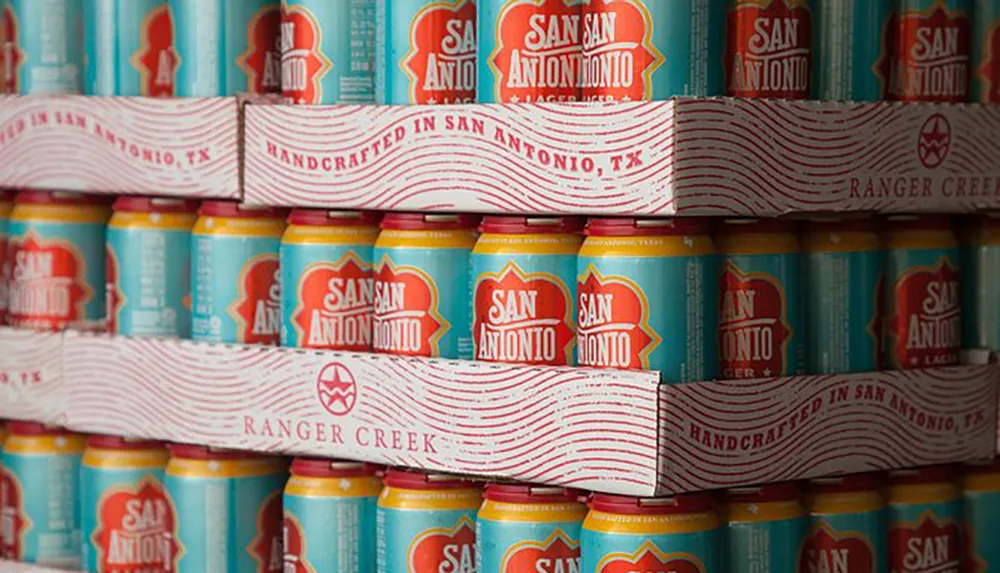 The image shows a stack of brightly colored cans of San Antonio Lager beer handcrafted in San Antonio TX by Ranger Creek