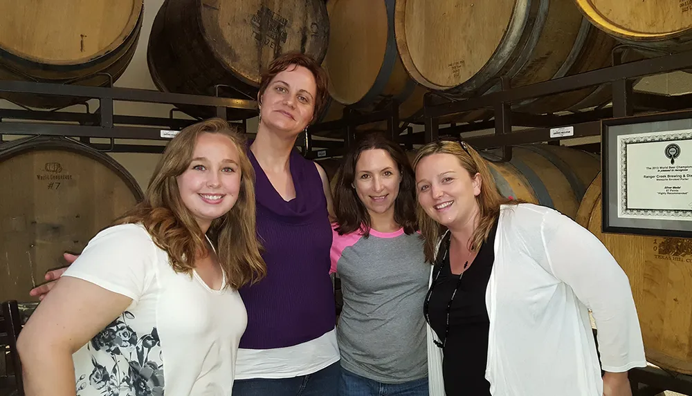 Four women are smiling for a photo in front of wooden barrels possibly at a winery or brewery