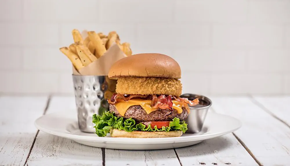 The image shows a gourmet burger with toppings and cheese accompanied by a side of seasoned fries and a dipping sauce presented on a white plate set against a white wooden background