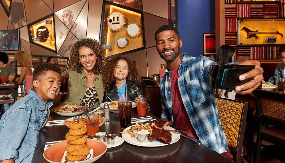 A family of four is smiling for a selfie while sitting at a restaurant table with various dishes in front of them
