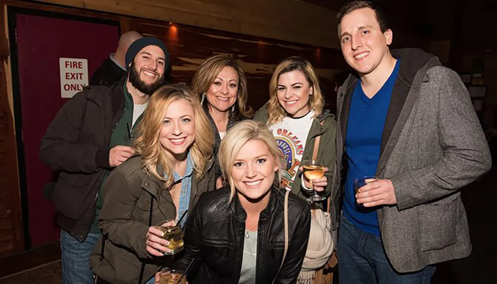 A group of six smiling people is posing for a photo indoors some holding drinks with a FIRE EXIT ONLY sign visible in the background