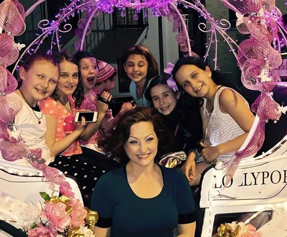 A group of happy children surrounds an adult woman posing for a photo with a whimsically decorated pink and purple themed background