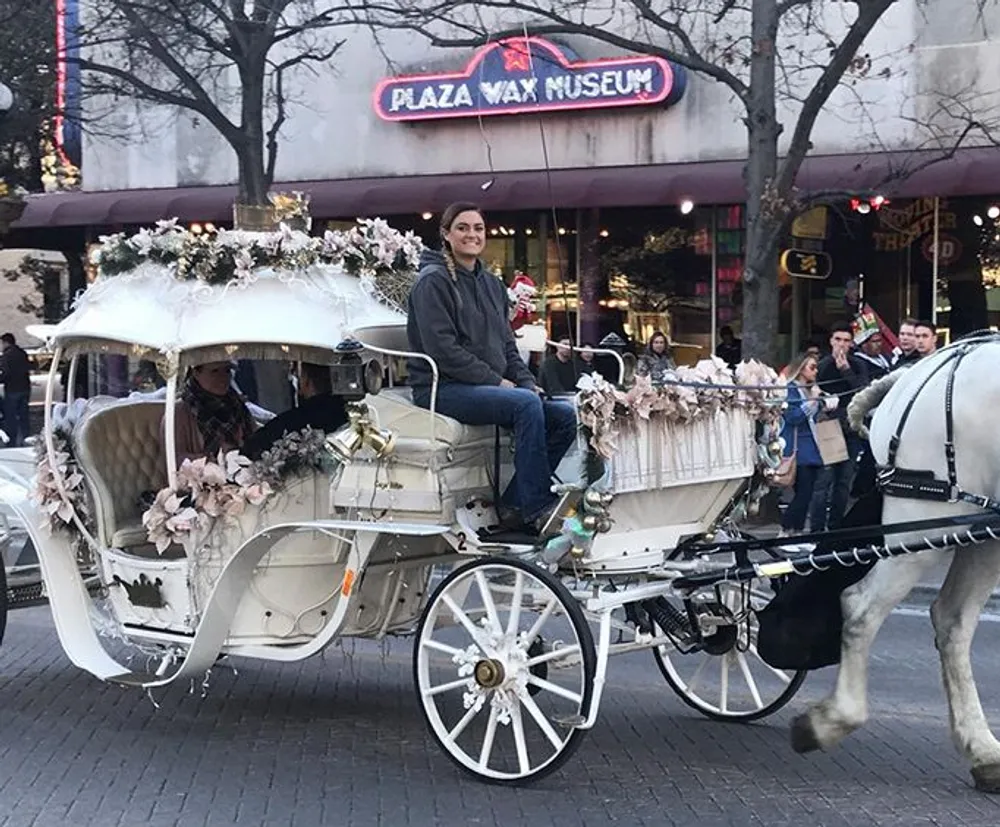 A person sits atop a festively decorated horse-drawn carriage in front of a wax museum smiling as passersby look on