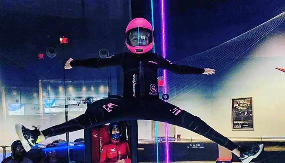 A person in a full-body suit and helmet is spreading their arms and legs while indoor skydiving suspended in mid-air by a powerful vertical wind tunnel with an instructor monitoring nearby