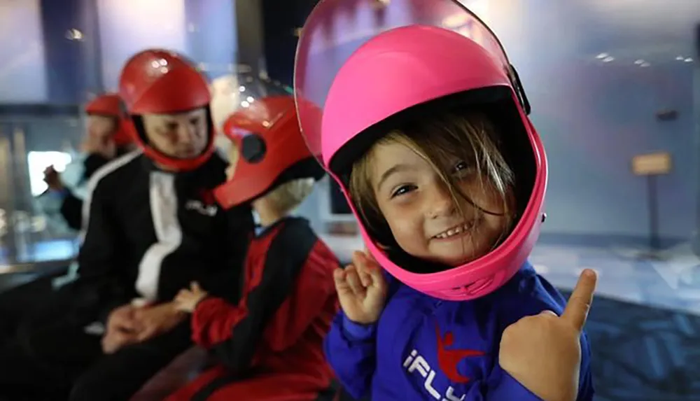 A child wearing a pink helmet gives a thumbs-up with a big smile likely excited for a simulated skydiving experience as others suit up in the background