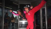 An instructor in a red suit guides a participant wearing a pink sleeve as they simulate skydiving in an indoor wind tunnel, with spectators watching from behind the glass.