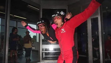 An instructor in a red suit guides a participant wearing a pink sleeve as they simulate skydiving in an indoor wind tunnel, with spectators watching from behind the glass.