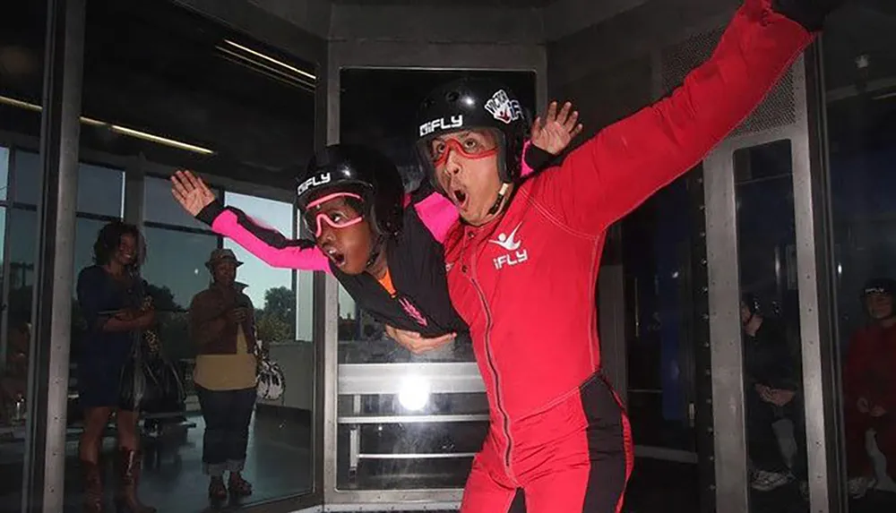 An instructor in a red suit guides a participant wearing a pink sleeve as they simulate skydiving in an indoor wind tunnel with spectators watching from behind the glass