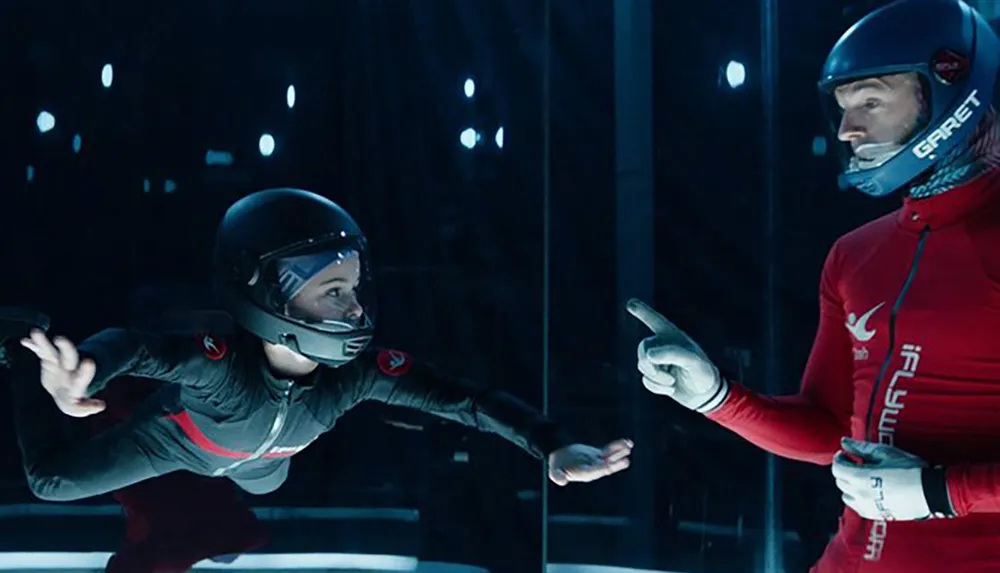 Two people in helmets and flight suits are practicing indoor skydiving guided by an instructor