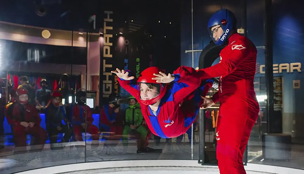 A person experiences indoor skydiving with an instructors assistance while others watch from behind a glass barrier