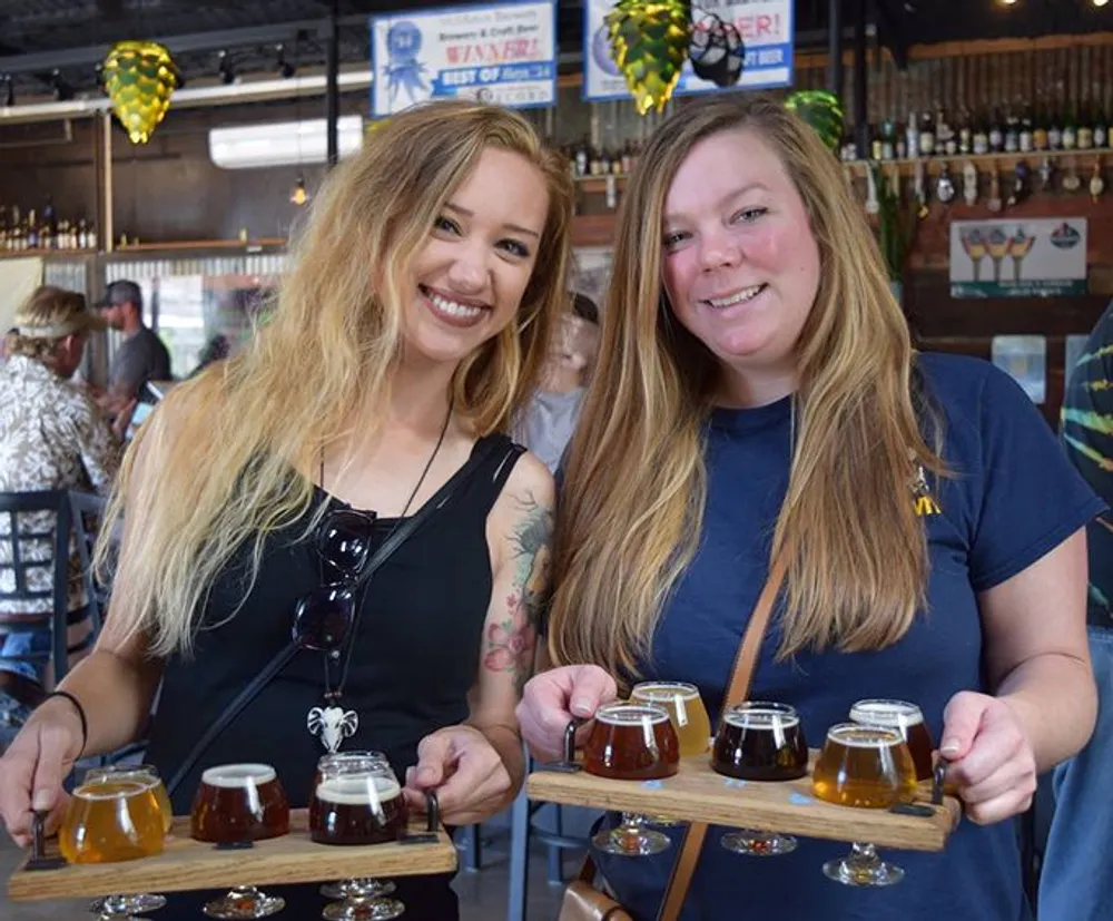 Two smiling women are holding wooden paddles with assorted glasses of beer likely enjoying a beer tasting experience at a brewery