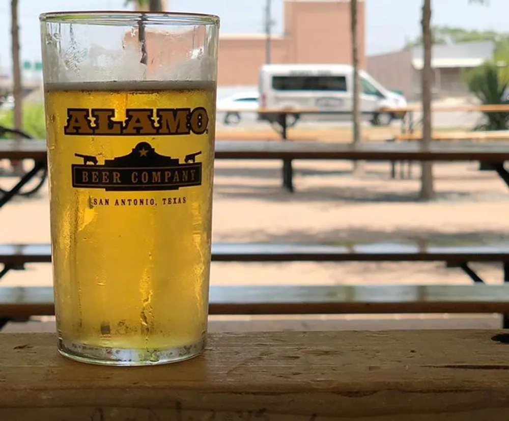 A half-full glass of beer from the Alamo Beer Company is resting on a wooden surface with an outdoor seating area in the background