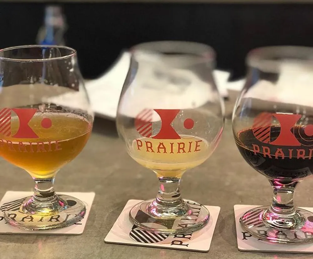 Three glasses of various beers are sitting on coasters on a bar each with the logo PRAIRIE printed on them