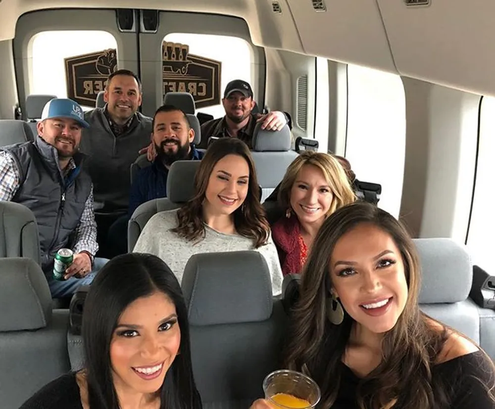 A group of smiling people is posing for a photo inside a vehicle with some holding drinks suggesting a social outing or celebration