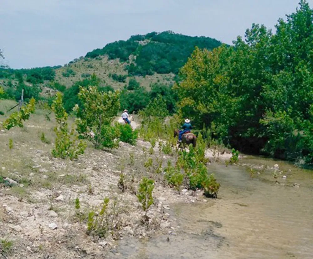 Two individuals are riding horses along a shallow stream that meanders through a lush green landscape