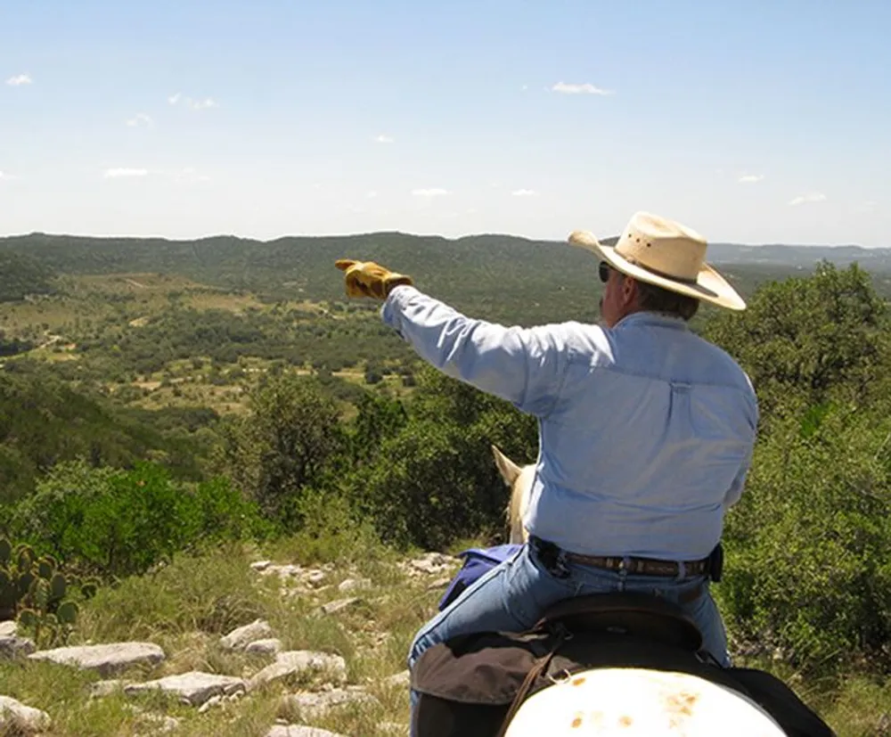 A person wearing a cowboy hat is pointing toward the horizon while sitting on a horse in a natural hilly landscape