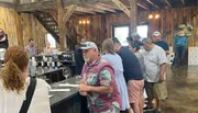 A group of people is gathered inside a rustic-styled building, possibly a winery or tasting room, with some waiting at the counter and others engaged in conversation.