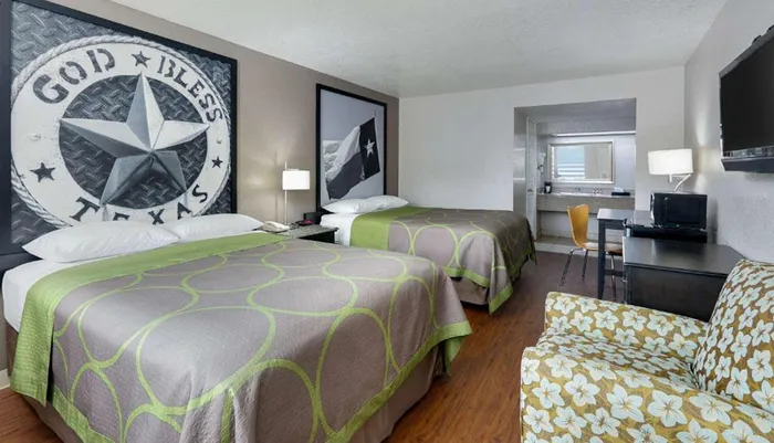 The image shows a hotel room with two beds featuring Texas-themed decor with a God Bless Texas wall emblem patterned bedspreads and a seating area