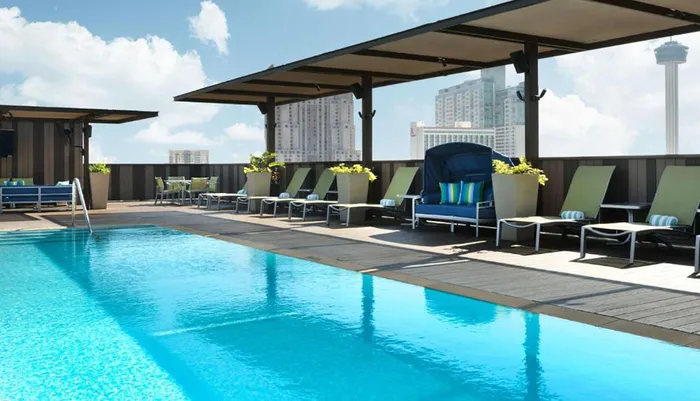 An outdoor rooftop swimming pool area with lounge chairs under shaded structures against a backdrop of high-rise buildings and a clear sky