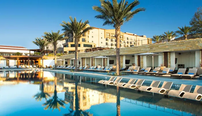 The image depicts a tranquil luxury resort pool flanked by sun loungers palm trees and a view of the hotels architecture under a clear blue sky