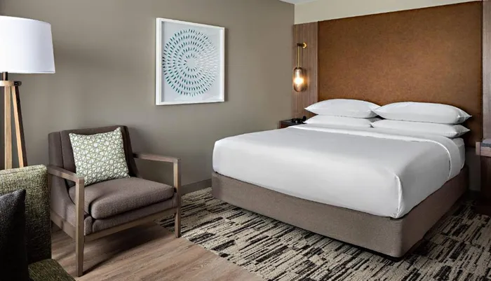 The image shows a neatly arranged modern hotel room with a large bed an armchair a floor lamp and decorative artwork on the wall