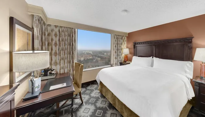 The image shows a well-appointed hotel room with a large bed a work desk and a city view through the window