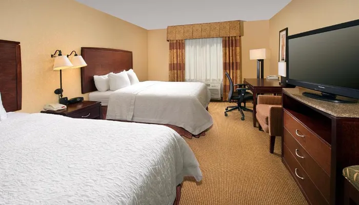 This is a neatly arranged hotel room with two queen-size beds a desk with a chair and a large flat-screen TV