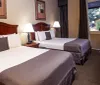 The image shows a neat hotel room with two beds nightstands with lamps framed artwork and a desk featuring a warm and inviting decor