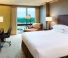 The image shows a neatly arranged hotel room with a large bed a work desk modern furnishings and a view of the cityscape through the window