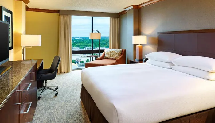 The image shows a neatly arranged hotel room with a large bed a work desk modern furnishings and a view of the cityscape through the window