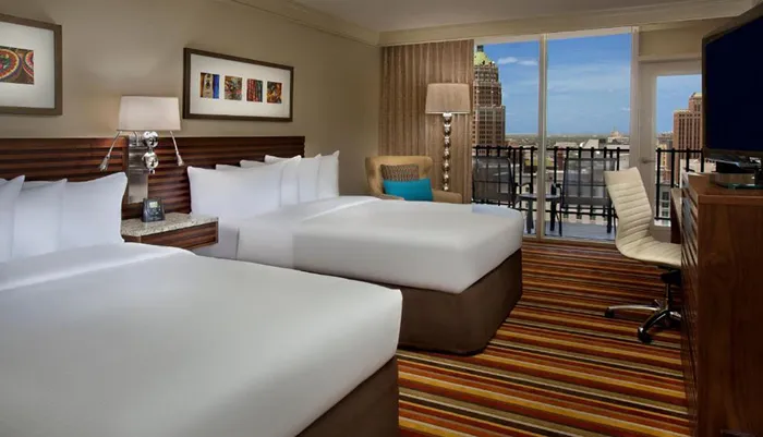 The image shows a modern well-appointed hotel room with two beds sleek furniture and a balcony that offers a city view