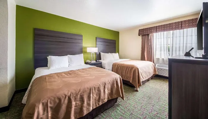The image shows a hotel room with two queen-sized beds patterned carpet a green accent wall and simple furnishings