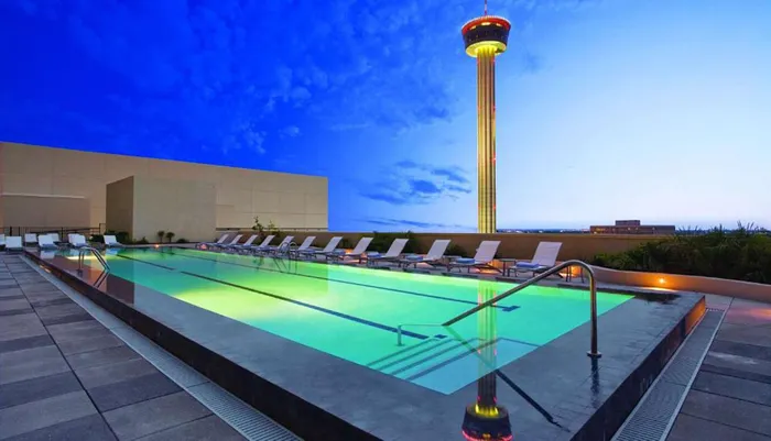 The image shows an outdoor swimming pool on a rooftop with lounge chairs and an illuminated tower in the background against a dusk sky