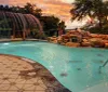 An outdoor swimming pool with a cascading rock waterfall and lit torches creates a tranquil atmosphere against a backdrop of a sunset-streaked sky