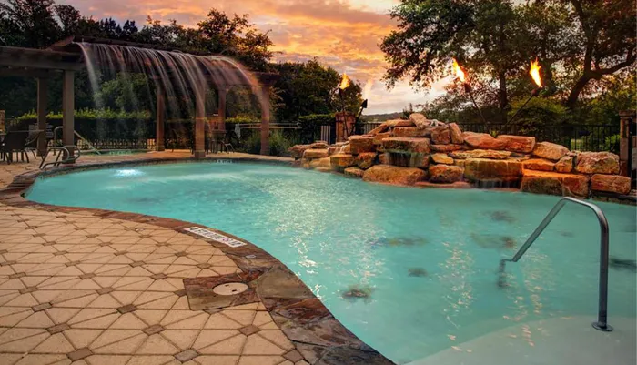 An outdoor swimming pool with a cascading rock waterfall and lit torches creates a tranquil atmosphere against a backdrop of a sunset-streaked sky