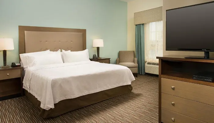 The image shows a neatly arranged hotel room featuring a large bed with white linen a flat-screen TV on a dresser a nightstand with a lamp and a sitting area by the window