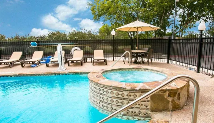 The image shows a sunny outdoor pool area with lounge chairs a hot tub and a patio set enclosed by a fence and surrounded by trees