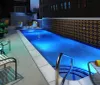 This image shows an illuminated outdoor swimming pool at nighttime with stylish poolside furniture and decorative tiles set against an urban building backdrop