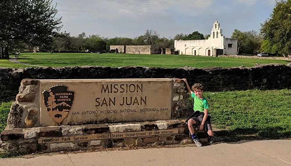 A young person is sitting on the edge of a stone sign for Mission San Juan at the San Antonio Missions National Historical Park