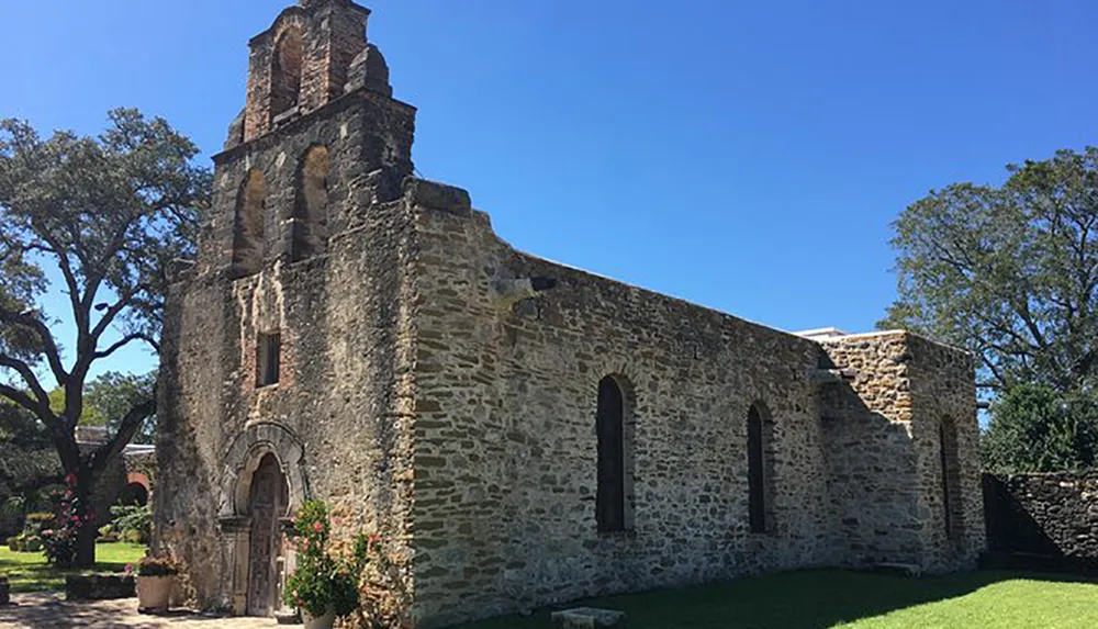 The image shows a well-preserved historic stone mission with an ornate bell tower under a clear blue sky