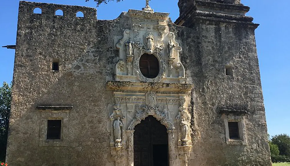 The image shows the faade of a historic stone mission with ornate carvings and an arched doorway under a clear blue sky