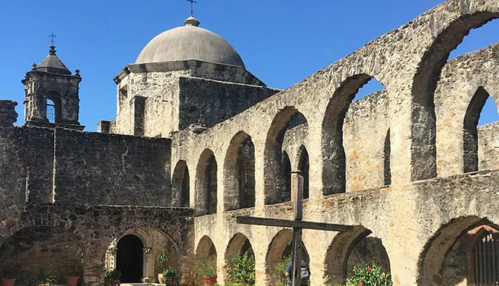 The image shows the historical architecture of an old stone mission with a dome and arches under a clear blue sky