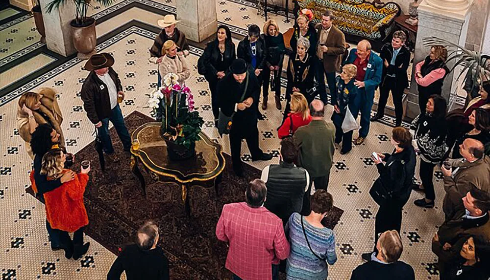 A group of people appears to be attentively listening to a person speaking in an elegant foyer with a black-and-white tiled floor