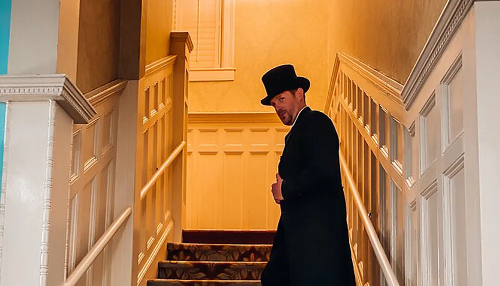 A man wearing a dark coat and top hat stands on a staircase looking back towards the camera