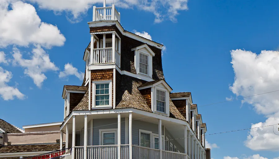 The image shows the upper part of a quirky architectural building with a sharply pitched roof and a widow's walk, contrasting with a bright blue sky with puffy clouds.