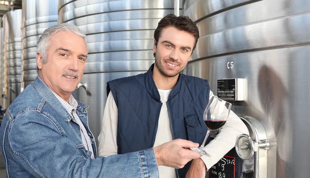 Two men one holding a glass of wine stand smiling in front of stainless steel fermentation tanks at a winery