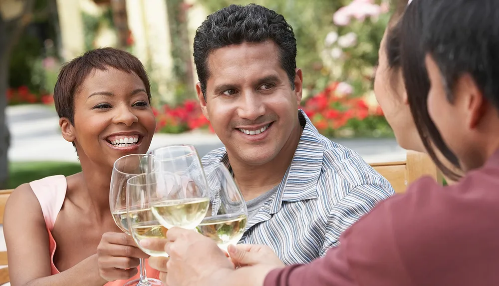 Three people are joyfully toasting with glasses of white wine sitting outdoors with flowers in the background