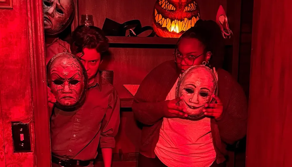 Two people illuminated by red light hold masks in front of their faces against a backdrop that includes a glowing pumpkin