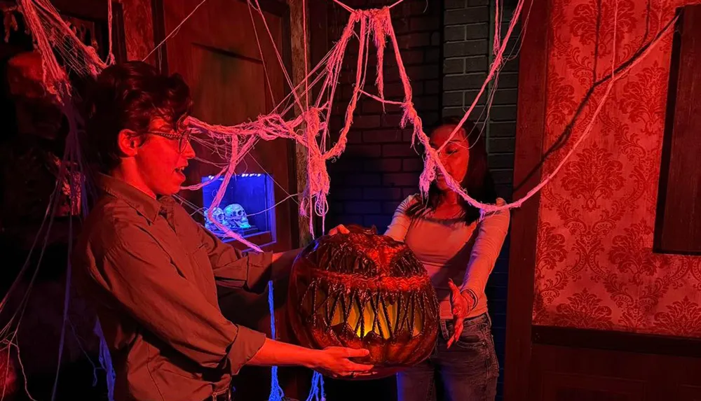 Two people in a dimly lit cobweb-filled room hold a large glowing pumpkin with a menacing carved face