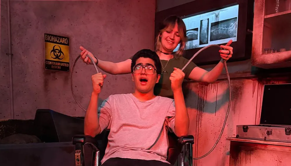 Two people are in a dimly lit biohazard-themed room with one sitting in a chair looking surprised and the other standing behind holding wires